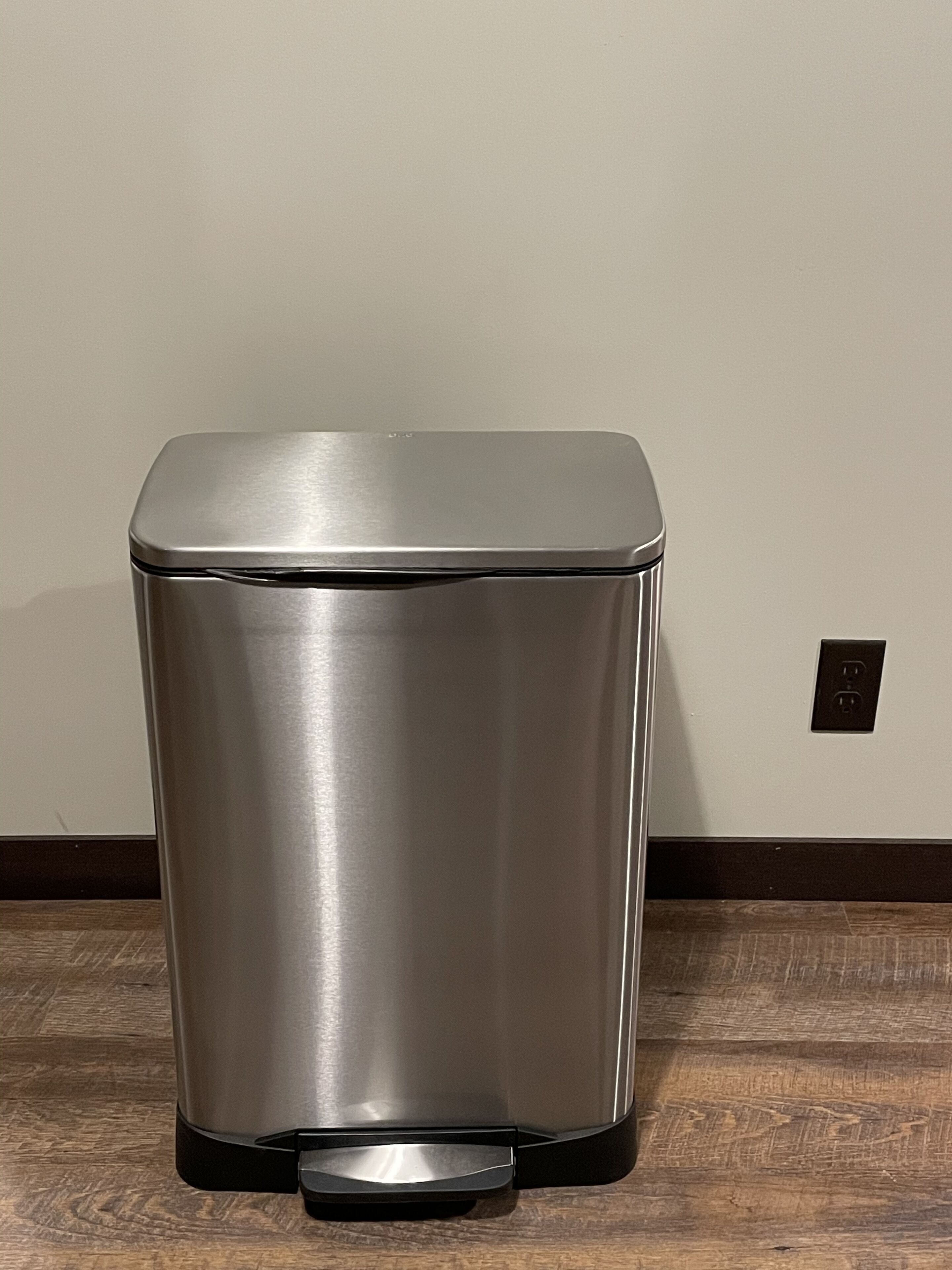 A stainless steel trash can with a black base, positioned against a plain wall with a visible electrical outlet.
