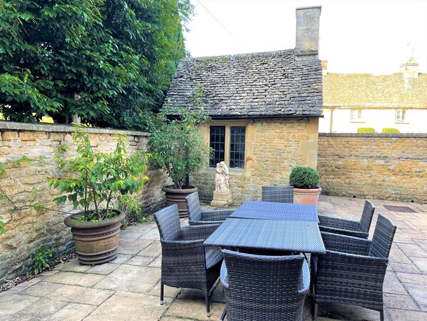 A stone cottage with a tiled roof, surrounded by potted plants and wicker furniture.