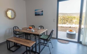 A dining area with a wooden table, four chairs, and a bench. There is a view of a landscape through a glass door.