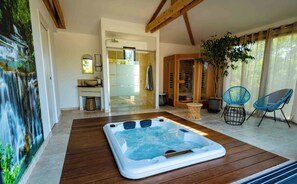 A spa area with a hot tub, wooden flooring, a large potted plant, and two blue chairs.