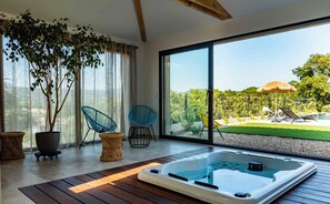 A modern room with a wooden floor, a large sliding glass door leading to a garden, a hot tub, and two blue chairs.