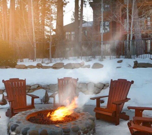 A stone fire pit with two wooden Adirondack chairs, surrounded by snow and a stone wall.