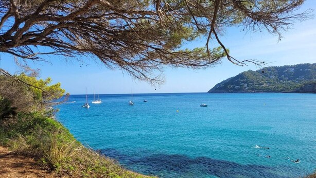 Paesaggio costiero con mare cristallino, imbarcazioni e colline sullo sfondo.