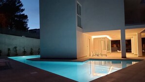 Un’area piscina esterna moderna con vasca limpida, zona relax coperta e un edificio con ampie vetrate.