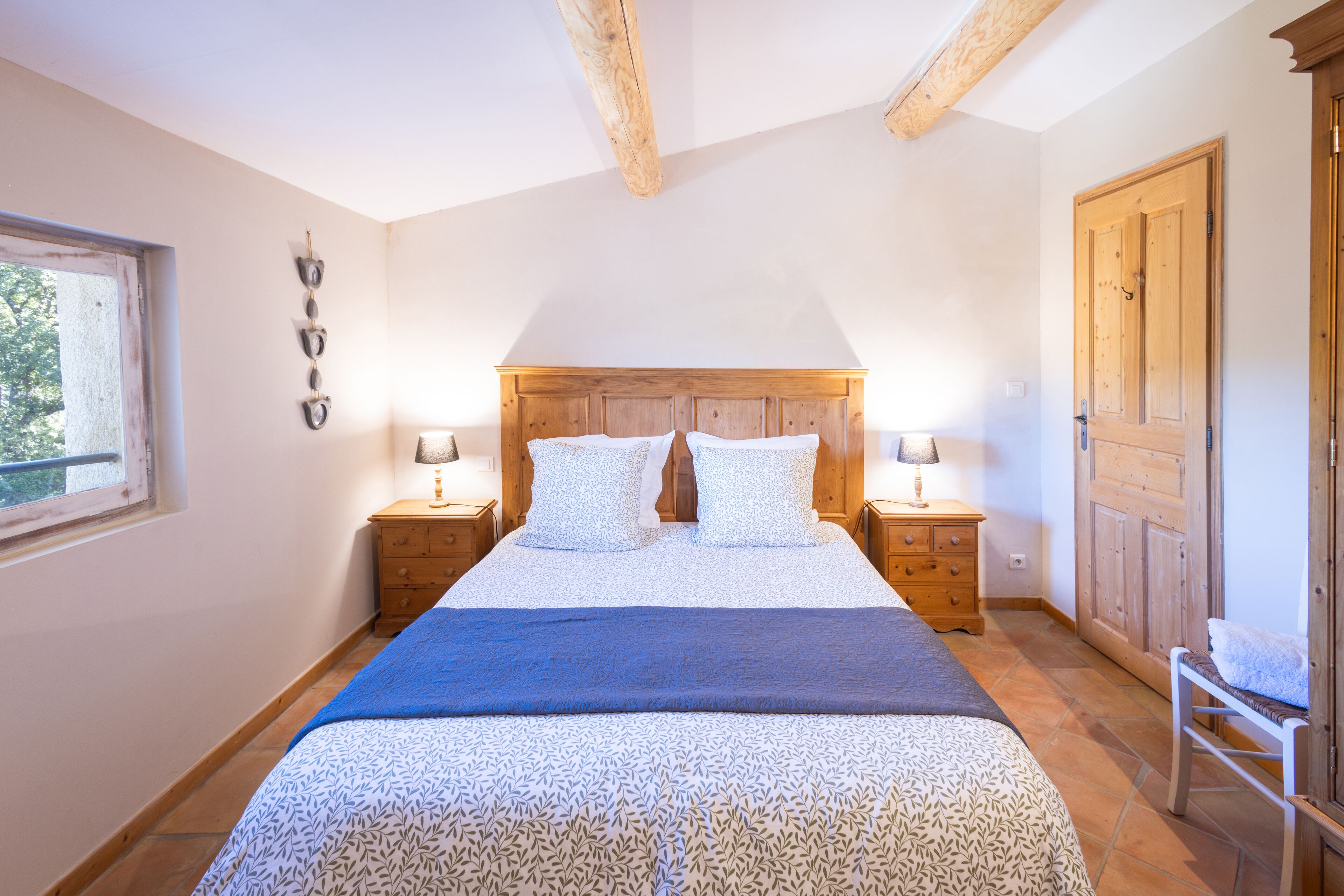 A bedroom with a large bed, wooden headboard, two bedside tables with lamps, a window, and a wooden door.