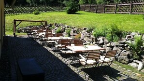 Un patio al aire libre con suelo de piedra, mesas y sillas de madera, rodeado por una valla de madera y vegetación.