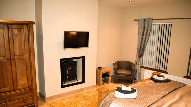 A bedroom with a wooden wardrobe, a flat-screen TV, a fireplace, a chair, and a bed with neatly folded towels. - Glenshane Country House (Maghera)