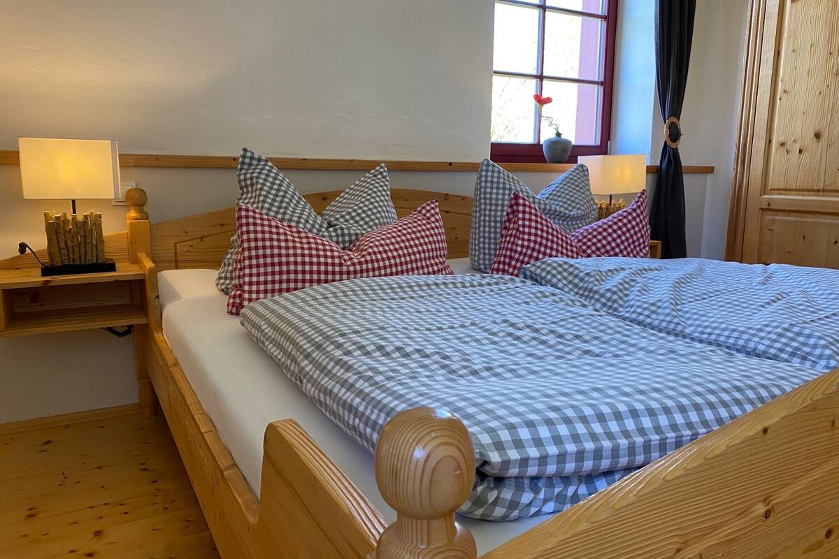 Un lit en bois avec une literie à carreaux, une table de chevet avec une lampe et une fenêtre avec des rideaux.