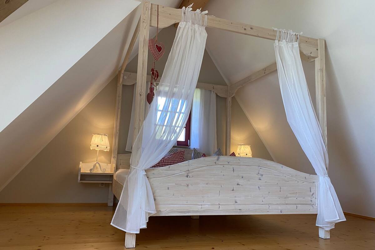 Un lit à baldaquin à quatre poteaux dans une chambre mansardée avec des poutres en bois, une table de chevet et deux lampes de chevet.