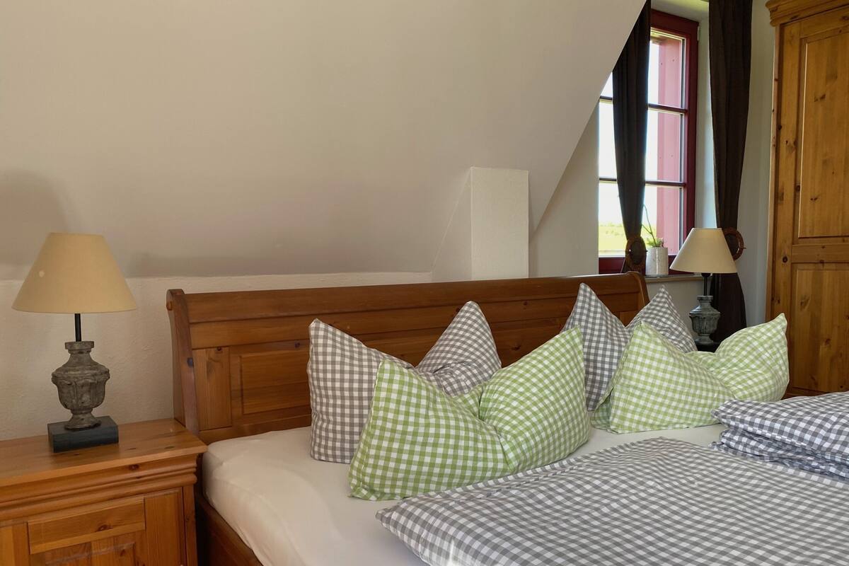 Une chambre  à coucher avec une tête de lit en bois, un lit recouvert d’une couette à carreaux, deux tables de chevet avec des lampes et une fenêtre avec des rideaux.