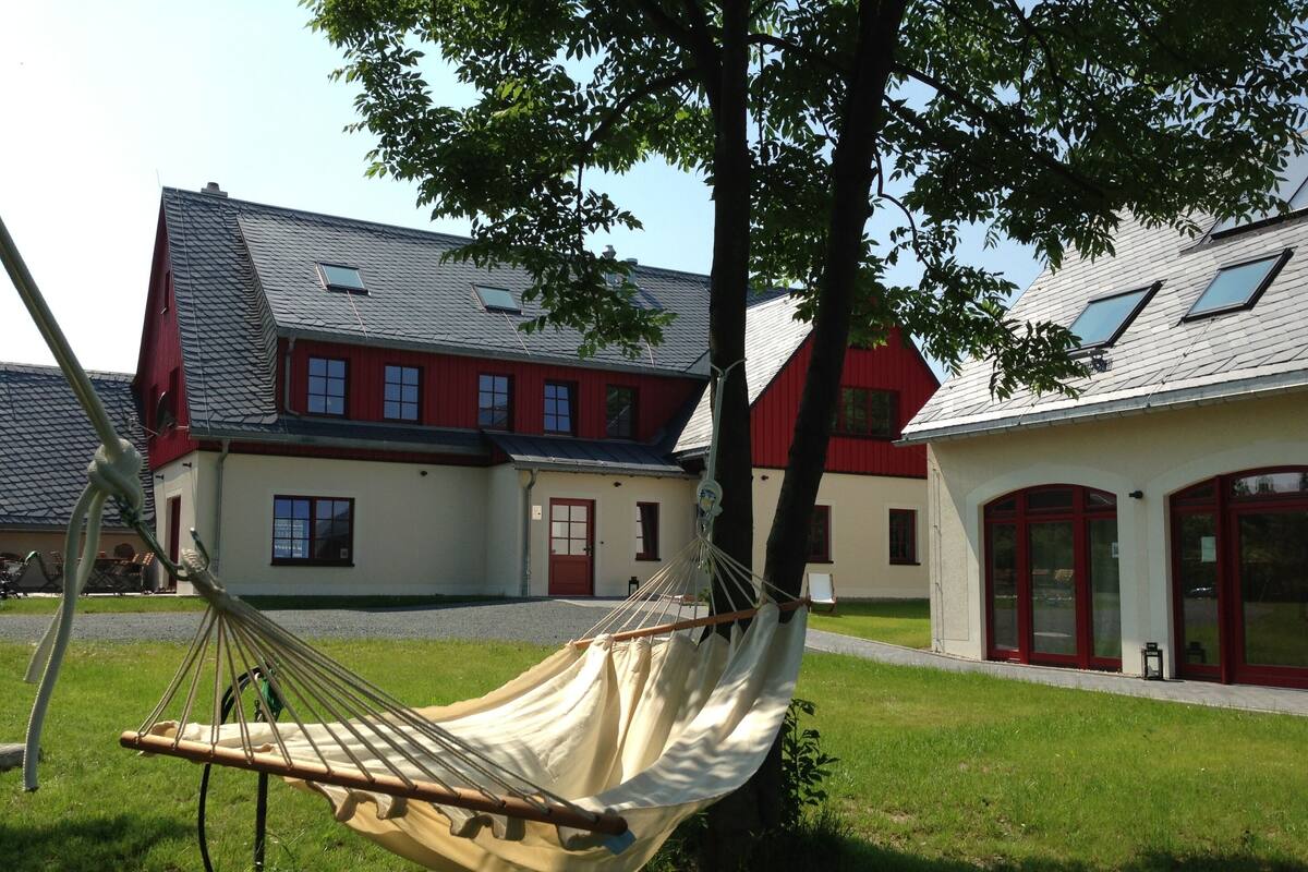 Un hamac est attaché à un arbre dans une zone herbeuse, avec un bâtiment en arrière-plan.