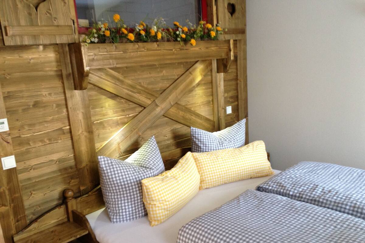 Un lit en bois avec une literie à carreaux et des oreillers, une tête de lit en bois et une petite étagère en bois ornée de fleurs.