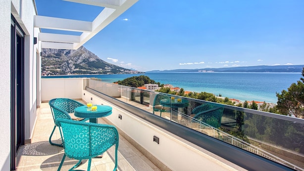 Ein Balkon mit Blick auf das Meer und die Berge, mit türkisfarbenen Stühlen und einem kleinen Tisch.