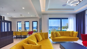 Ein modernes Wohnzimmer mit gelben Sofas, einem schwarzen Couchtisch und einem Essbereich mit gelben Stühlen. Große Fenster bieten Blick auf das Meer und die Berge.