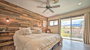 A bedroom with a wooden wall, a bed with white bedding, a ceiling fan, and a large window with a view of the outdoors. - Desert Oasis with Pool, Fire Pit, Game Room, and Dry Bar (Borrego Springs)