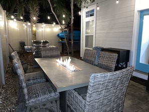 A patio with a fire pit table, wicker chairs, a grill, and string lights.