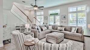 A spacious living room with a sectional sofa, a round ottoman, and a striped armchair. A staircase with a wooden railing leads to another level. - Family Beach Escape! Pool Access + Kayaks, Bikes & Paddleboard Included (Santa Rosa Beach)