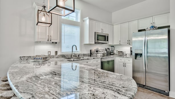 A modern kitchen with white cabinetry, a stainless steel refrigerator, a granite island, and pendant lighting. - Family Beach Escape! Pool Access + Kayaks, Bikes & Paddleboard Included (Santa Rosa Beach)