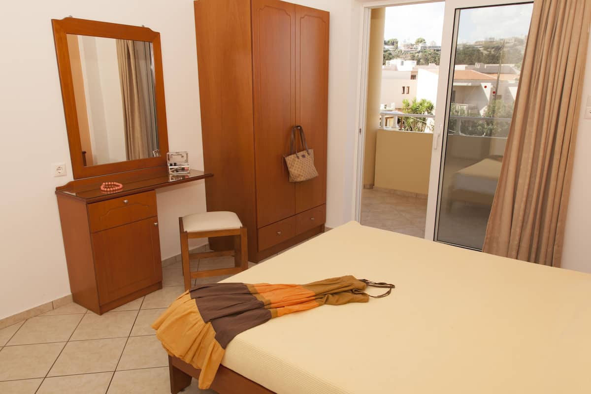 A bedroom with a bed, a wooden wardrobe, a small stool, a mirror, and a view of the outside balcony.