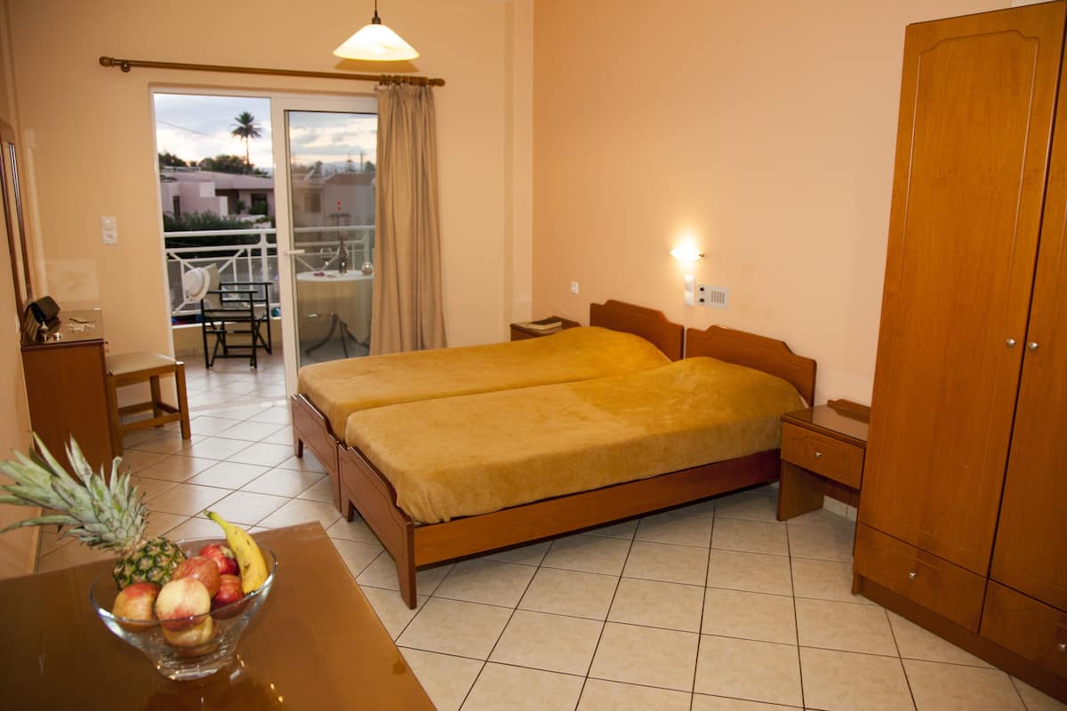 A hotel room with two beds, a dining table with fruit, a balcony with a chair, and a wardrobe.