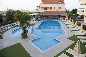 Una piscina con sillones de descanso y sombrillas, rodeada de jardines y un edificio con tejado de tejas rojas.