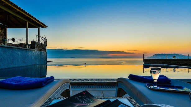 A poolside lounge with a view of the sunset over the water, lounge chairs, and a glass of water. - Infinite Blue Epidavros (Palaia Epidavros)