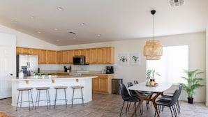A modern kitchen with wooden cabinets, a central island with stools, a dining table with chairs, and a hanging light fixture.