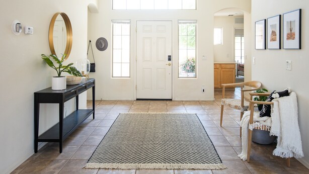 A clean, modern entryway with a large white door, a console table with a potted plant, a black rug, and a wooden bench.