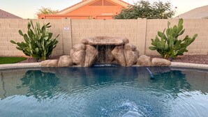 A swimming pool with a rock waterfall feature, surrounded by cacti and a brick wall.