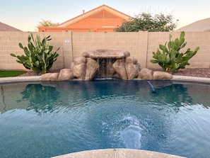 A swimming pool with a rock waterfall feature, surrounded by cacti and a brick wall.
