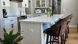 Uma cozinha moderna com armários brancos, uma ilha central com banquetas, micro-ondas e máquina de café.