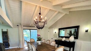 Uma sala de jantar com mesa de madeira, cadeiras brancas, um lustre e uma televisão sobre um móvel.