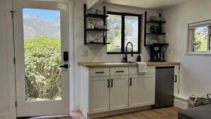 Cozinha moderna com armários brancos, bancada de madeira e lava-louças embutida. Há uma janela com vista para vegetação e montanhas.