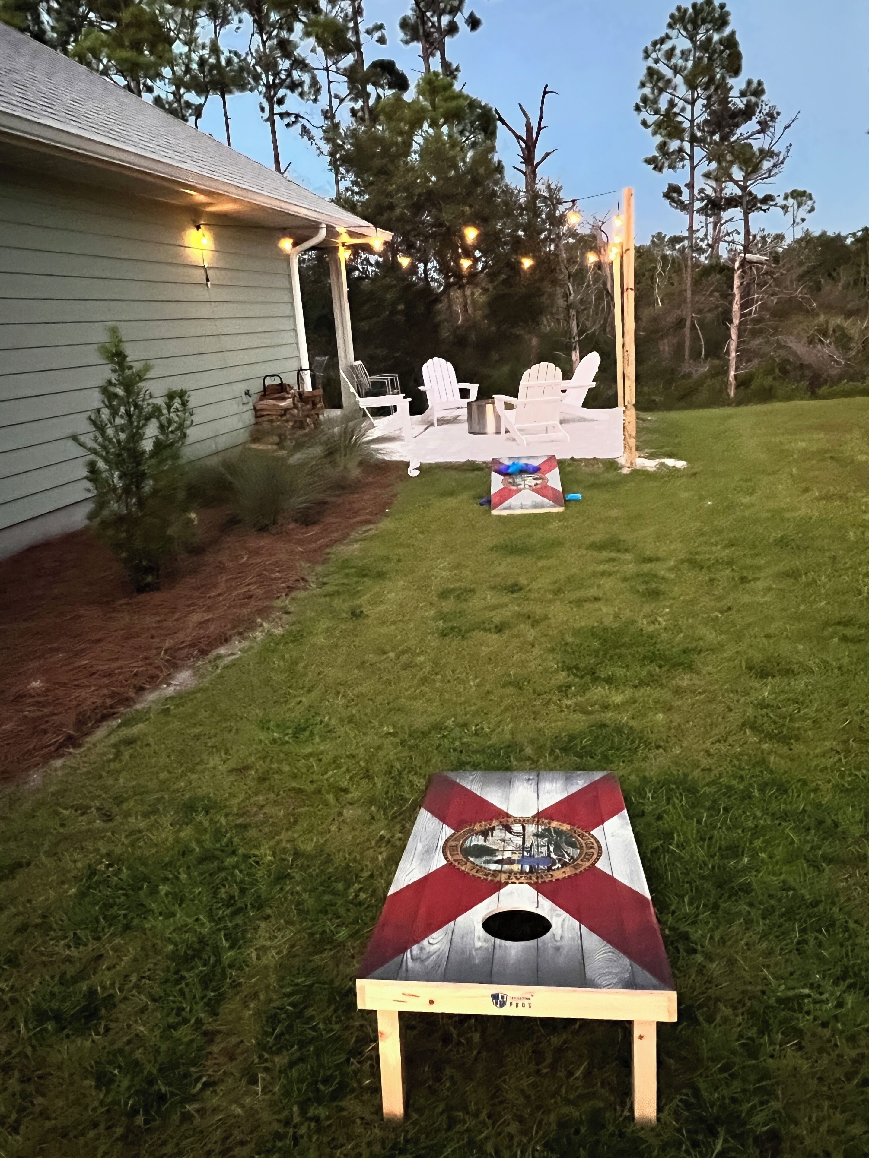 Un pati amb un tauler de cornhole de fusta, cadires blanques i un arbre petit.