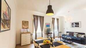 En moderne stue med sofa, spisebord og fjernsyn. Gardiner, en lysekrone og billedkunst på væggene.