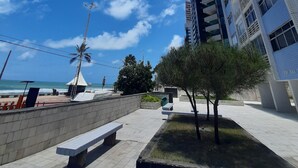A high-rise building with a clear blue sky and a few clouds. There is a bench and a small tree in the foreground. - Furnished apartment on Avenida Boa Viagem, beachfront condominium. (Recife)
