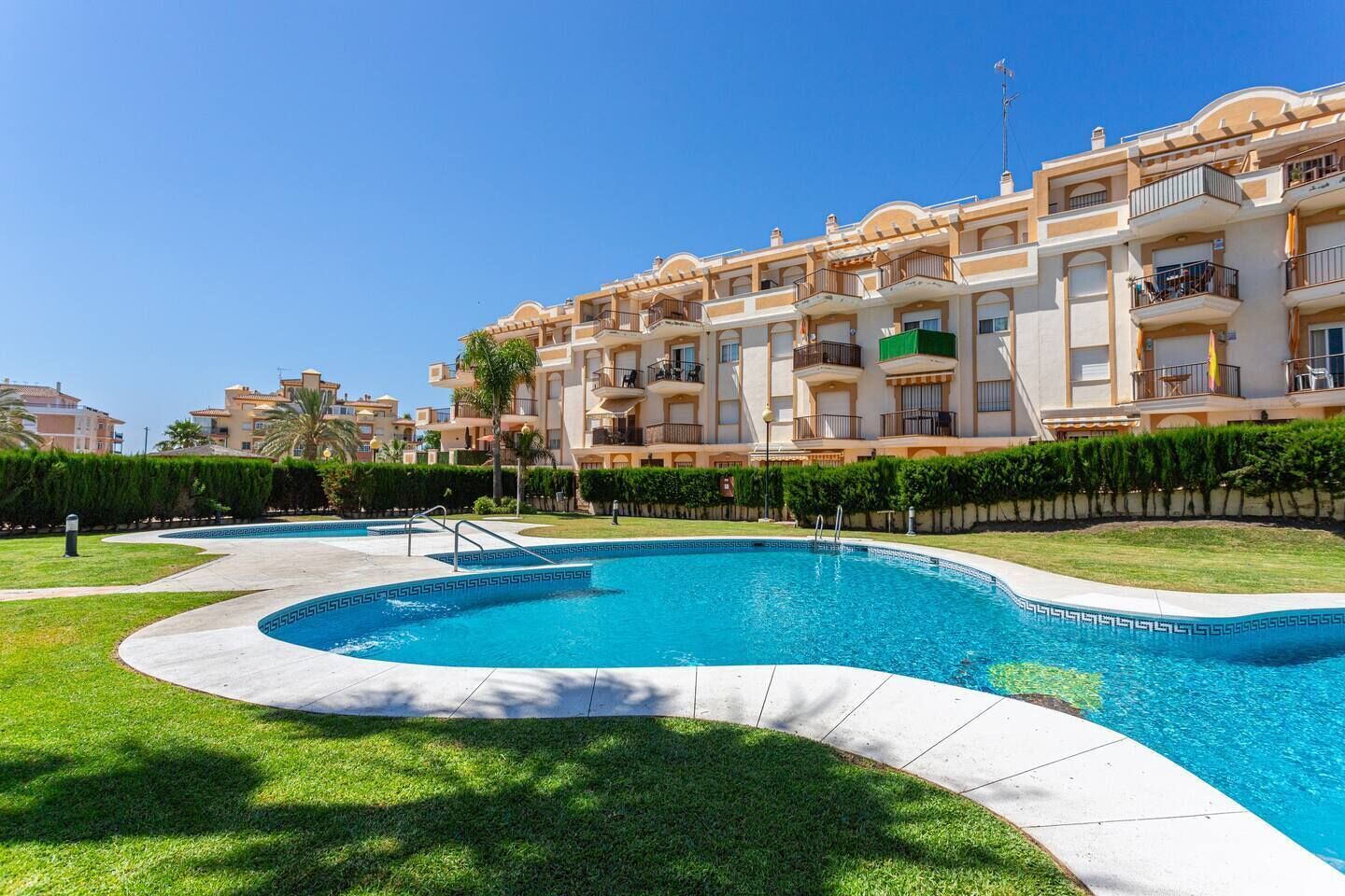A swimming pool with a curved design, surrounded by a well-maintained lawn and a row of multi-story buildings with balconies.