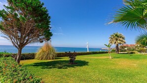Een verzorgde tuin met verschillende bomen en struiken, een helder uitzicht op zee, en een wit gebouw op de achtergrond.