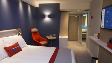A hotel room with a bed, a red chair, a small table with a bottle, a mirror, and a screen displaying room information.