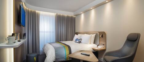 A hotel room with a bed, bedside table, chair, and a small desk with a remote control.