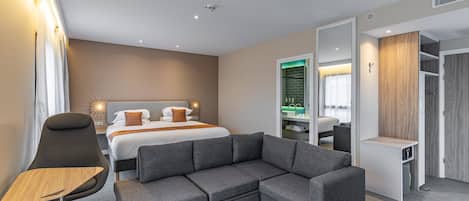 Une chambre d’hôtel moderne avec un lit, un canapé gris, une table basse ronde et un dressing.