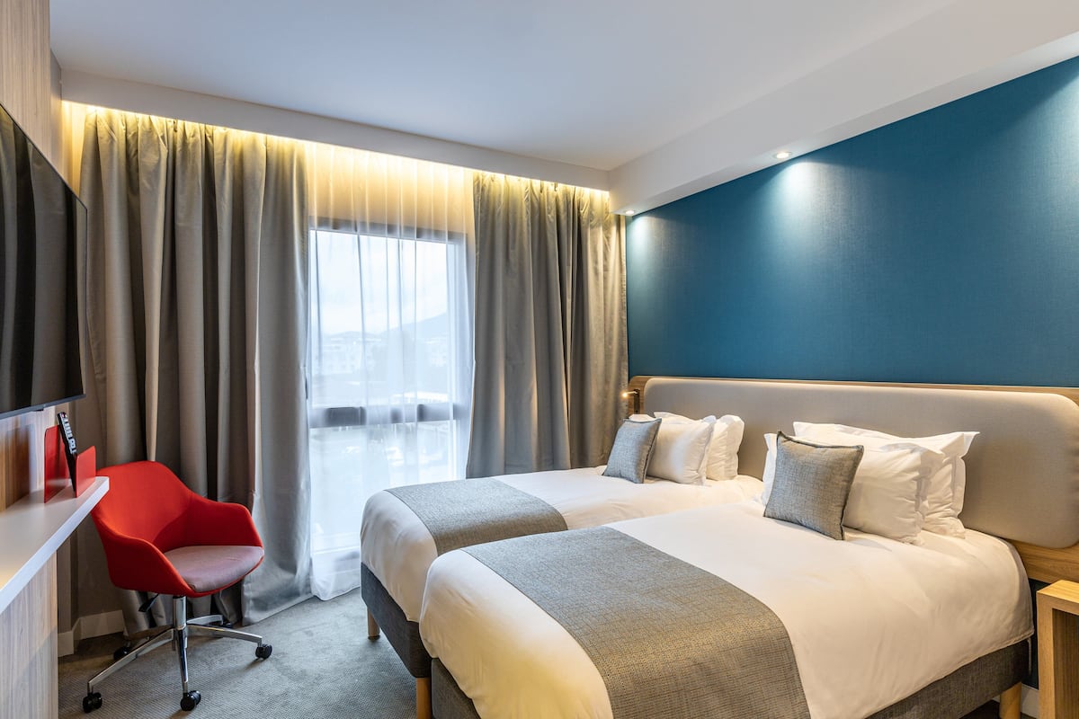 A hotel room with two beds, a desk with a red chair, and a large window with curtains.