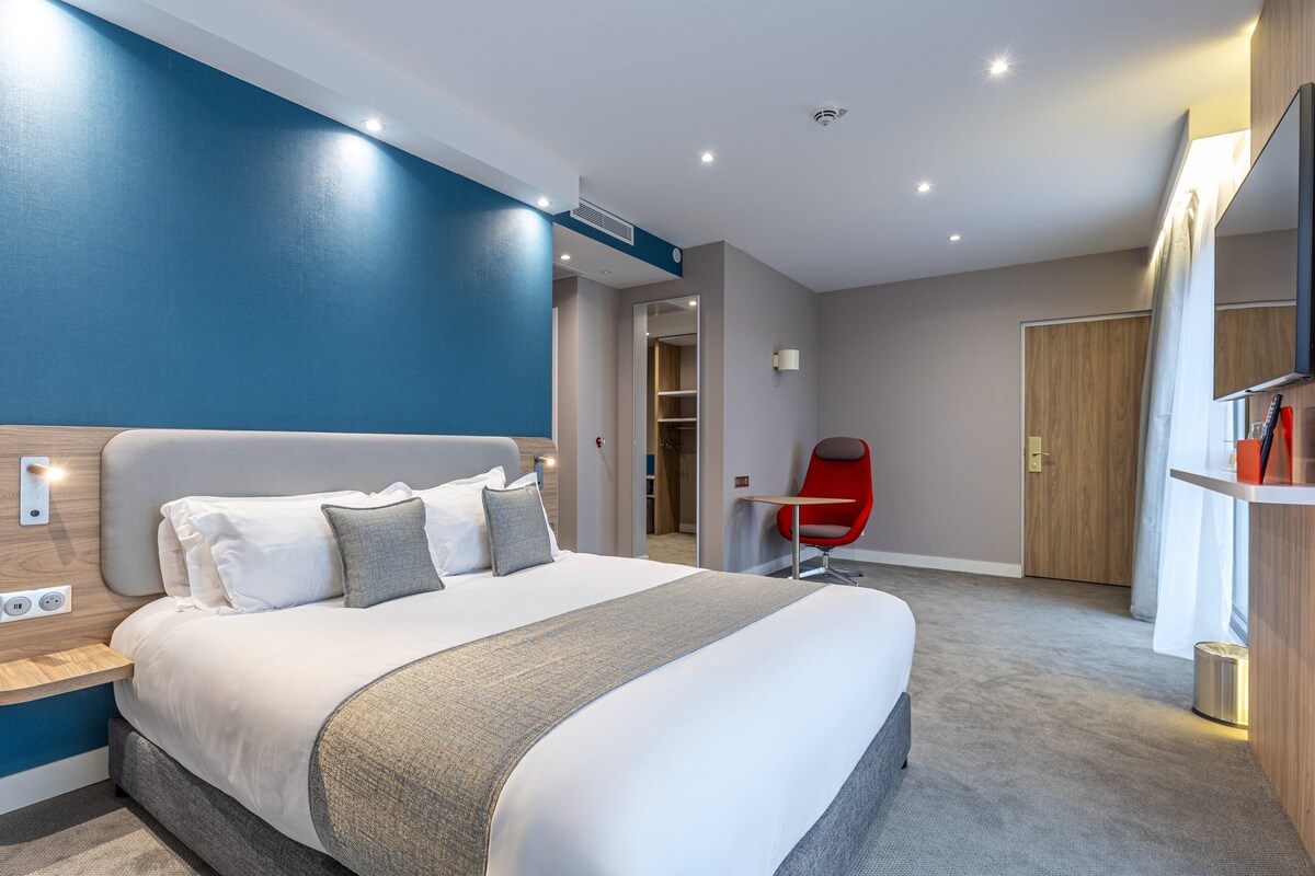 A modern hotel room with a large bed, a red chair, and a wooden wardrobe.