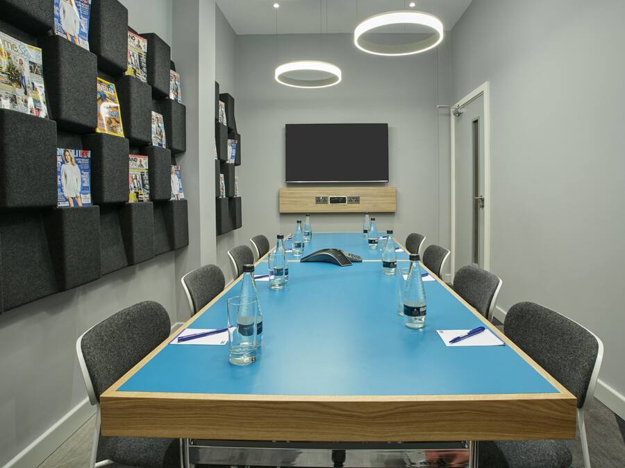 Un salón de conferencias con una larga mesa, sillas, botellas de agua y revistas en estanterías.