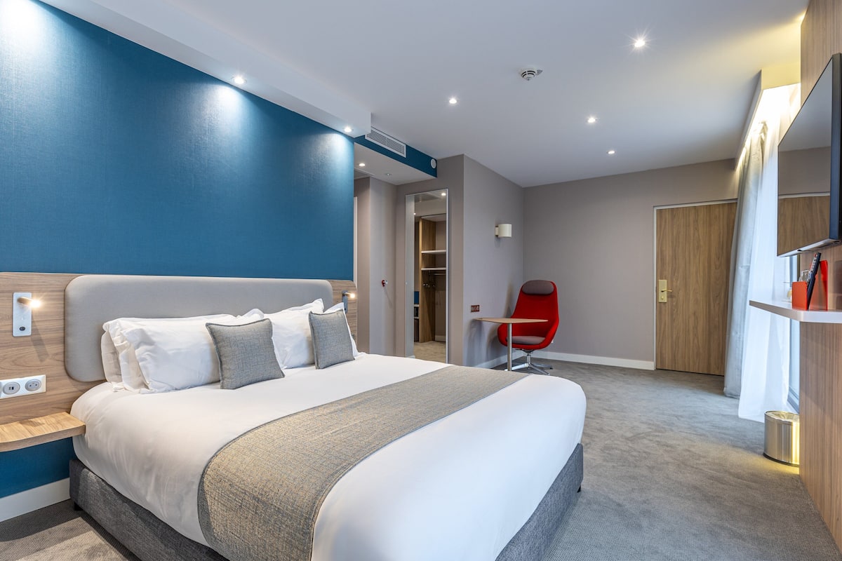 A modern hotel room with a large bed, a red chair, and a wooden wardrobe.