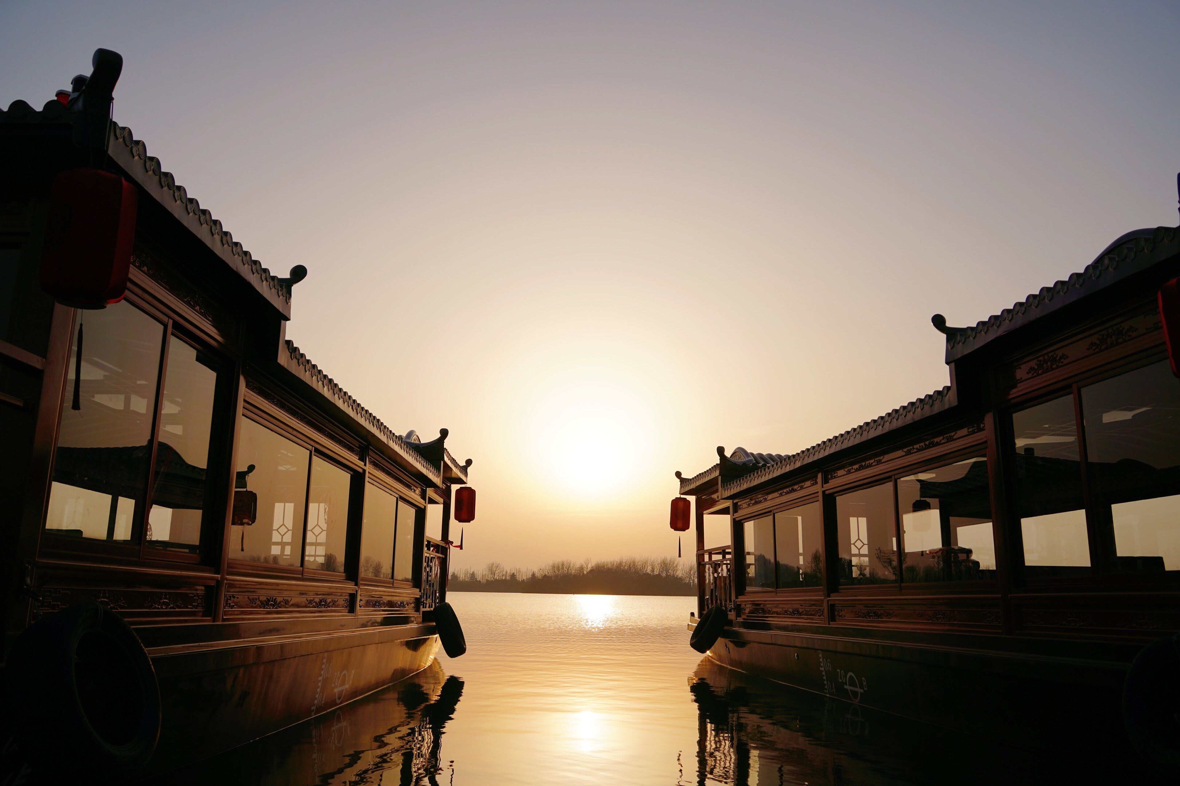 A traditional boat with a wooden structure and windows, docked at a calm body of water during sunset.