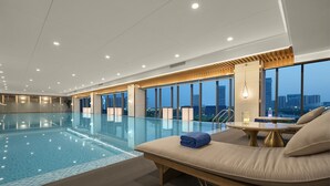 A modern indoor swimming pool with a lounge area, large windows, and a city view.