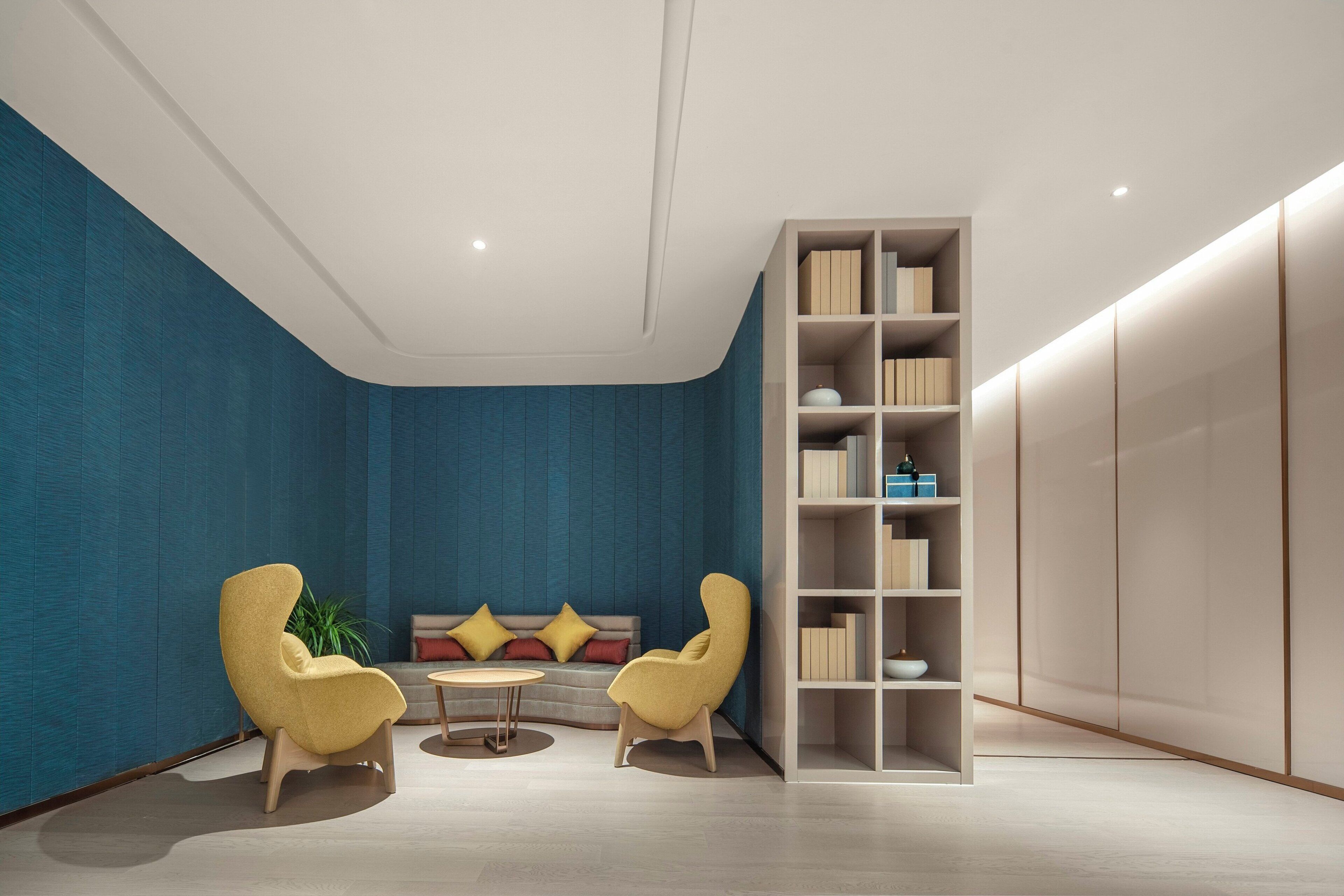 A modern living room with a blue accent wall, two yellow armchairs, a round coffee table, a sofa with cushions, and a built-in bookshelf.