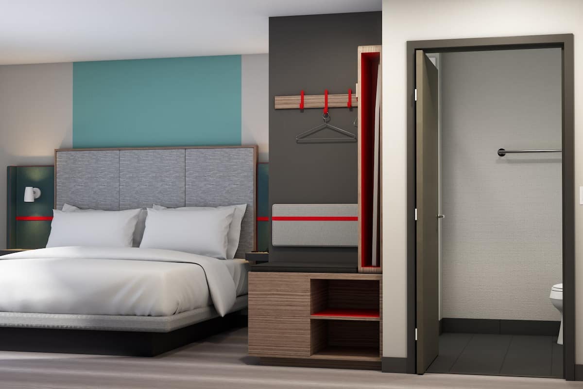 A modern hotel room with a bed, a wardrobe with a red accent, and a bathroom visible through an open door.