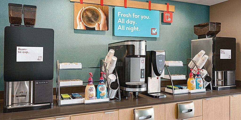 Una estación de café con varias máquinas de café, un letrero que dice “Recién hecho para ti. Todo el día. Toda la noche.” y un molinillo de café de pared.